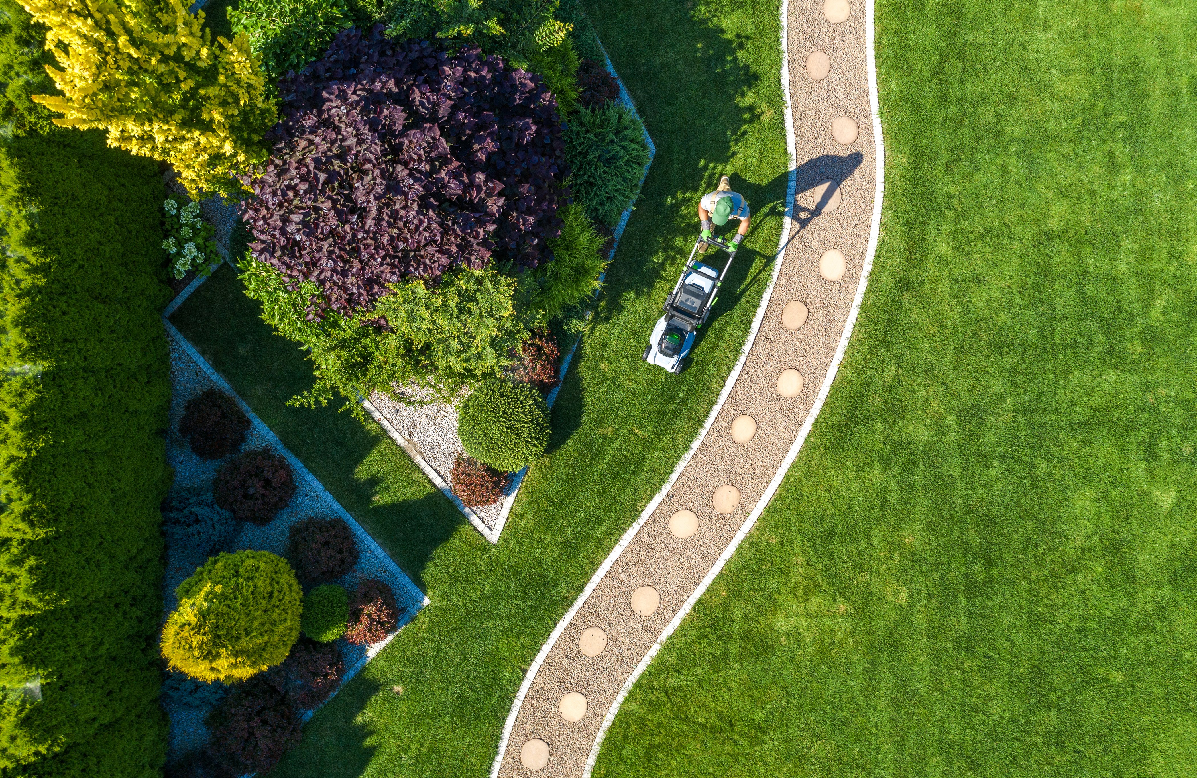 Large Beautiful Residential Backyard Garden Grass Mowing Aerial View. Caucasian Gardener Pushing Electric Cordless Grass Mower and Trimming the Lawn.